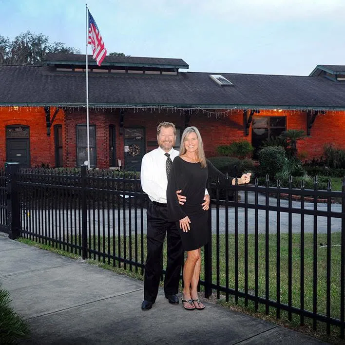 A man in a white shirt and tie and a woman in a black dress stand by a black iron fence in front of a red brick building.